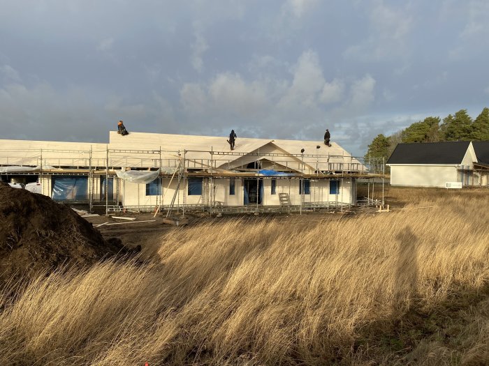 Hus under konstruktion med arbetare på taket och byggställningar runt fasaden.