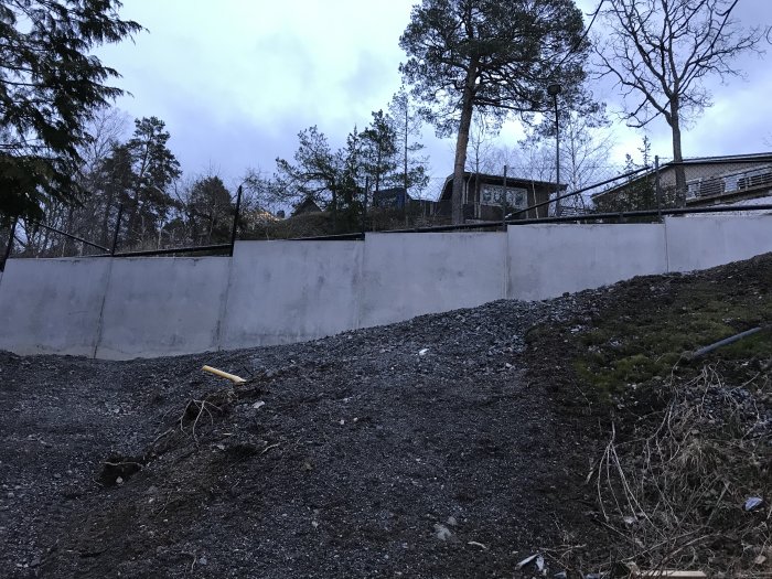 Nybyggd grå betongstödmur med grus och kuperad terräng framför, träd och hus i bakgrunden.