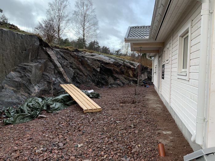 En ofärdig innegård vid en nybyggd vit husfasad med sprängsten och höga bergskanter i sluttningstomt.