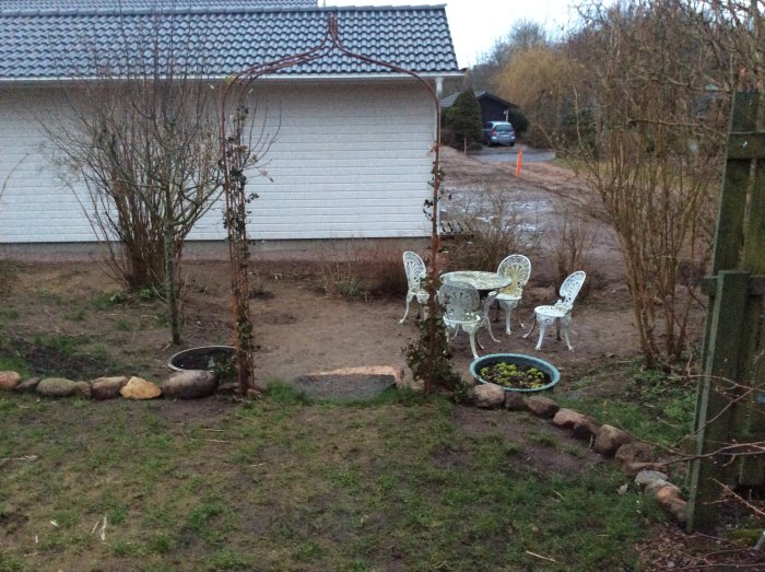 Trädgårdshörna med stentrappa, gräsmatta, vita trädgårdsmöbler och garagevägg i bakgrunden.