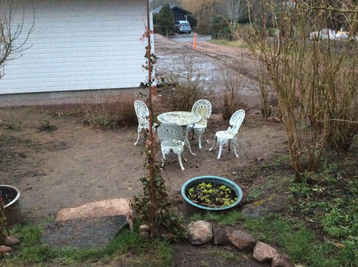 Oanlagd trädgårdshörna med en stenlagd trappa, metallmöbel, väggen till ett vitt garage, planteringskärl med grönt och nakna buskar.
