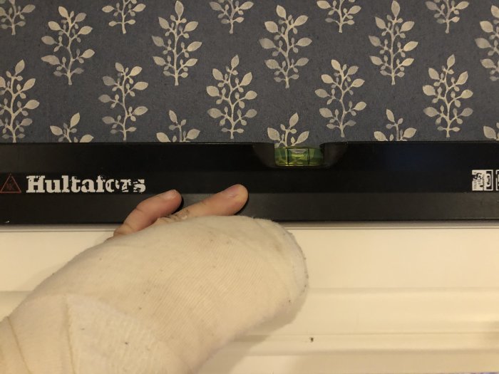 Hand i gips med tumregeln som använder vattenpass från Hultafors mot blommig tapet.