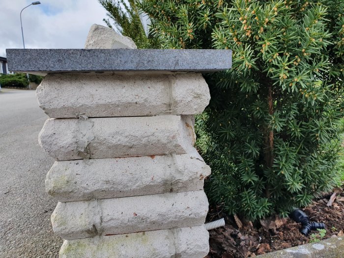 Stapelade betongblock med en flat sten ovanpå intill en grön buske.