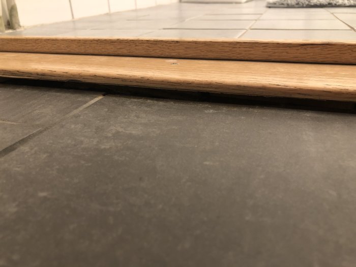 Trätröskel ovanpå grå klinkergolv med synliga mellanrum och underliggande träbitar.