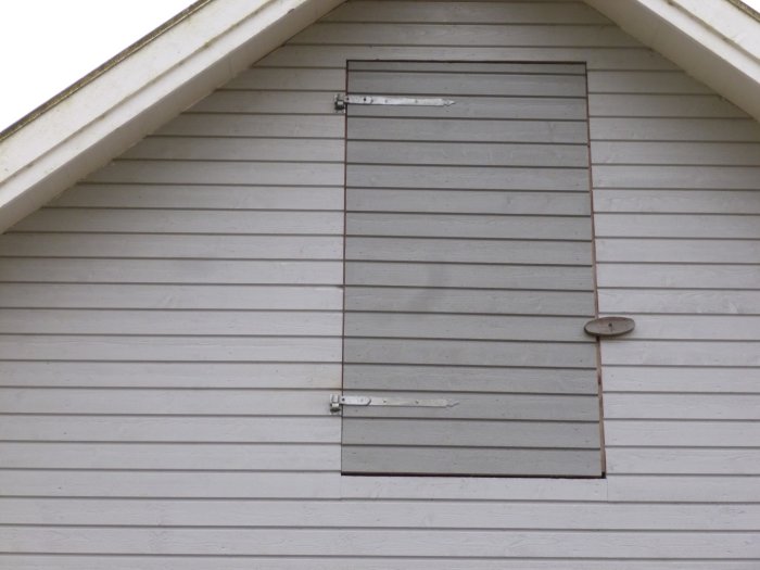 Lucka på en vit trävägg med krökt gångjärn, trolig vindlucka till stallvind, utan synligt läckage.