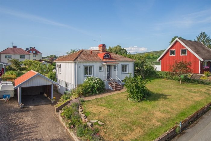 Vit 30-talshus med rött tak och spröjs i takkupan, carport till vänster, grön trädgård och rött hus i bakgrunden.