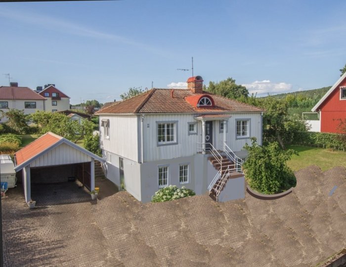 30-talshus med vita väggar och röd takkupa från ovan, angränsande carport, grönska runtomkring.