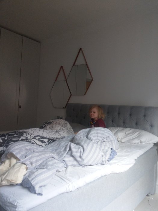 Barn sitter på sängkant i sovrum och ser på, oordnat täcke och kuddar, dekorativ väggspegel i bakgrunden.