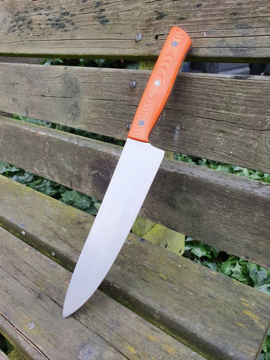 En kniv med orange micarta-handtag och svart G10-mellanlägg mot en träbänk.