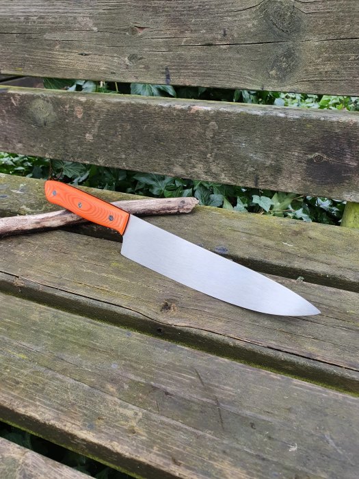 Handgjord kniv med orange micartahandtag och svart G10 mellanlägg på träbänk.