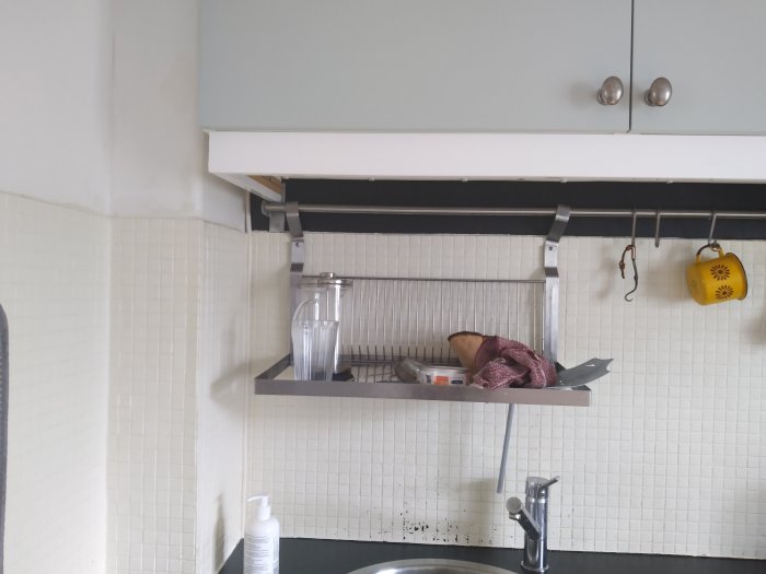 Kök med vit mosaikvägg, skåp och en hylla med köksredskap ovanför diskho.