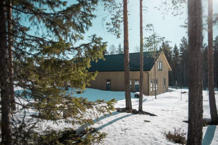 Träbeklätt hus med mörkt tak i snöig skogsmiljö under klarblå himmel.