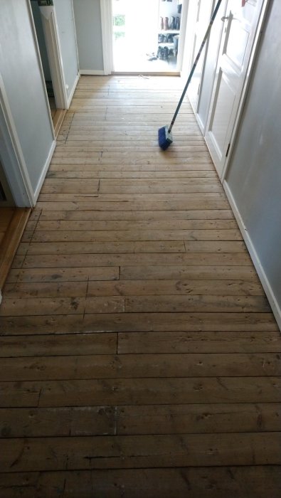 Gammalt plankgolv i en korridor med synliga spikar och slitage, en sopkvast står till höger.