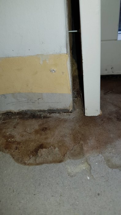 Fuktfläckar på betonggolv och skada på sockel vid hörn inomhus indikerar vattenskada.