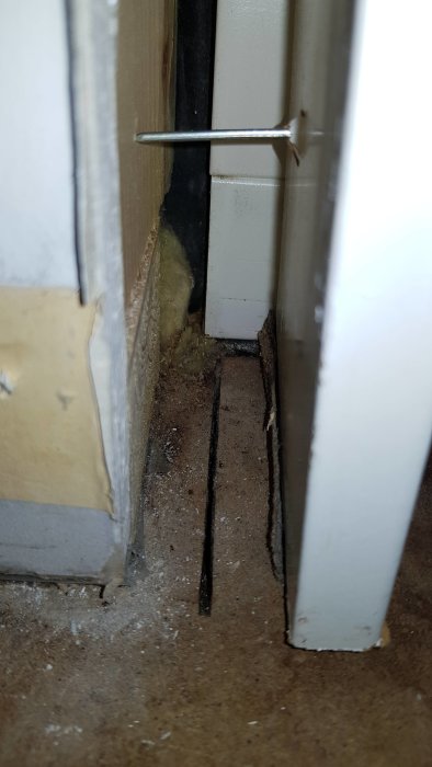 Vy av en inre hörn av ett rum som visar golv och väggbeklädnad, med synlig isolering och ett glapp.