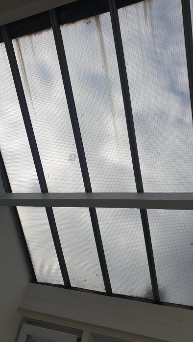 Vy uppåt genom ett befintligt tak med genomskinliga glaspaneler och en synlig träramstruktur.