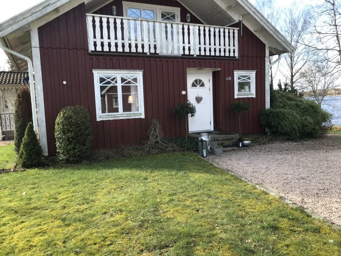 Rödmålat trähus med vit dörr och fönstertrim, veranda på övre våning, grön gräsmatta och utsikt över en sjö.