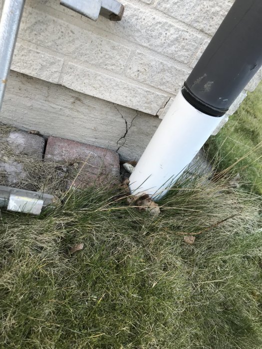 Spricka vid hörnet av en betongsockel nära ett stuprör och murad vägg, omgiven av gräs och enstaka tegelstenar.