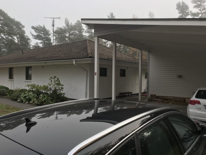 Enstaka hus med carport och vit fasad sedd från gård med del av bil i förgrunden.