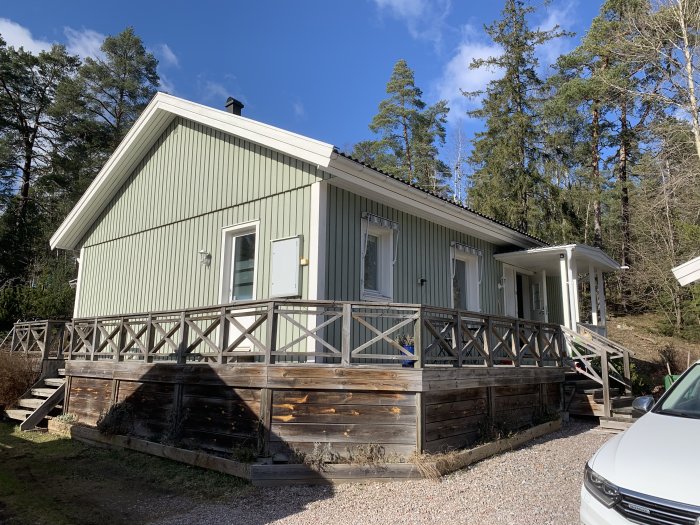 Ett grönmålat hus med vitmålade fönsterkarmar, försedd med en träterrass och omgivet av tallskog under solig himmel.