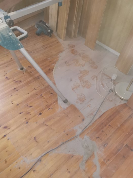 Slipdamm och fotspår på ett trägolv bredvid en stege och en slipmaskin under en renovering.