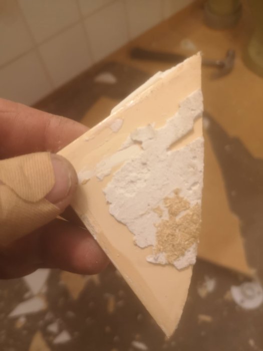 Någons hand håller en triangelbit av ett beige mattmaterial med vita och beige fläckar som kan vara lim eller underliggande skikt.