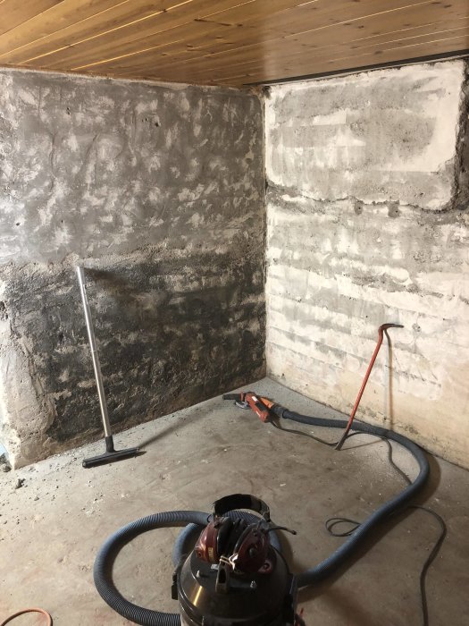 Rivningsarbete inuti hus, synligt sliten betongvägg, dammsugare och verktyg på betonggolv under renovering.