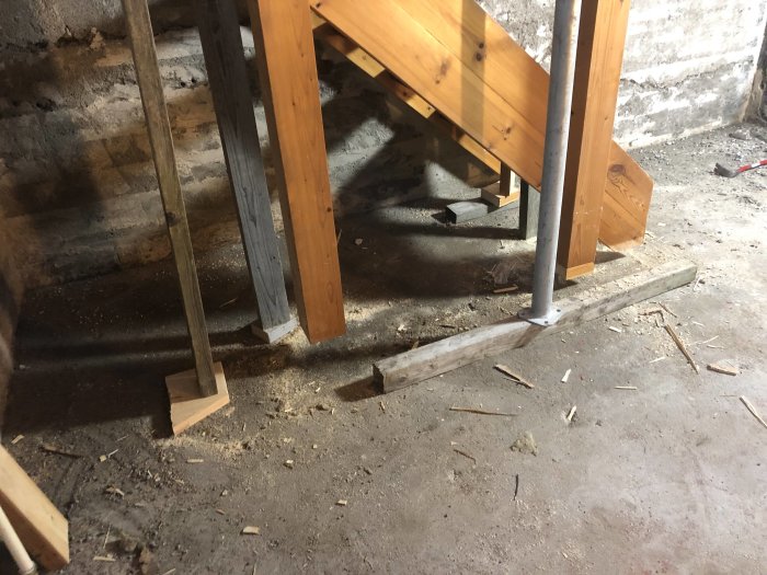 Rivningsavfall och träspån på ett betonggolv med synliga fundament, vid en delvis nedmonterad trätrappa.