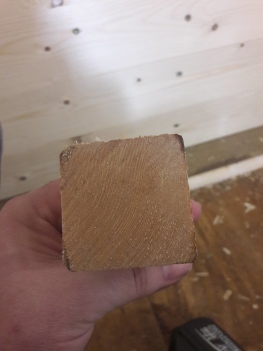 Hand håller upp en nyköpt 45x45 regel med synlig träådring mot en ljus träbakgrund.