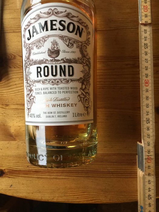 En Jameson-whiskyflaska på ett träbord bredvid en måttstock som visar centimeter.