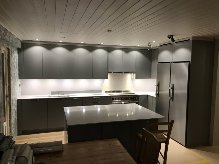 Modernt kök utan synliga uttag, gråa skåp, vita bänkskivor och integrerad belysning.