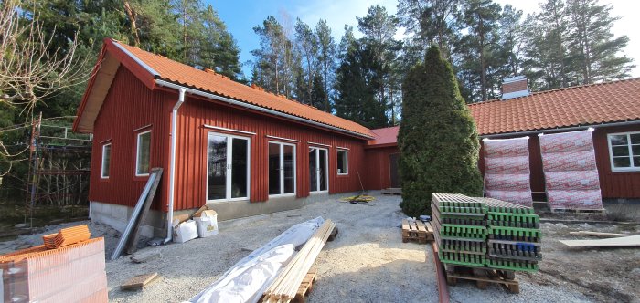 Rödfärgat hus under renovering med nya fönster, dörrar och delvis lagt tegel, byggmaterial framför huset, i soligt väder.