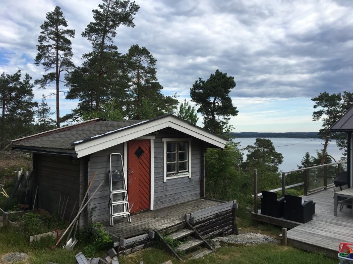 Nybyggd friggebod vid havskanten omgiven av tallar med klar himmel i bakgrunden, intill terrass och byggmaterial.