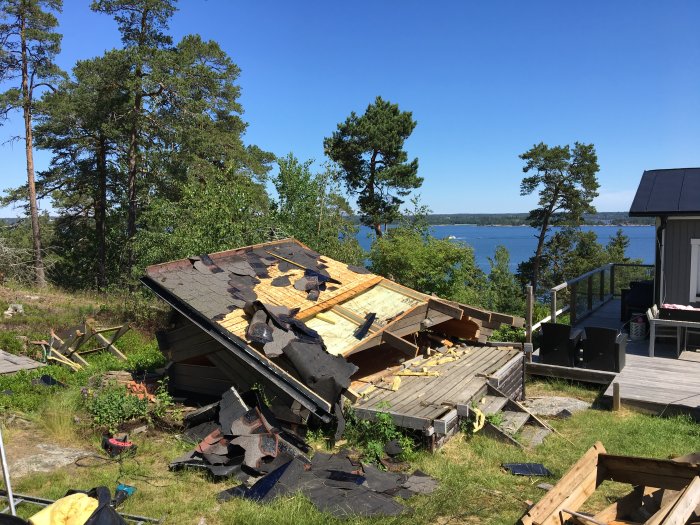 Nerriven gammal friggebod med spillror på marken, vid havsutsikt och gröna träd i bakgrunden.