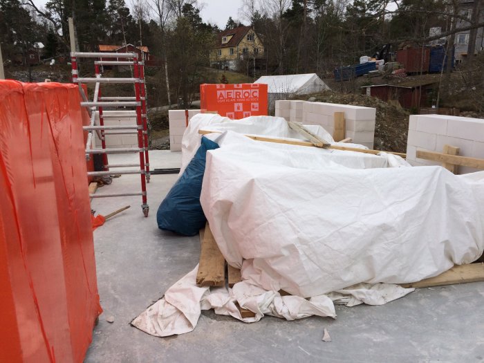 Byggplats med vit täckta murblock och byggmaterial inför murning av tredje våningen, ställning i bakgrunden.