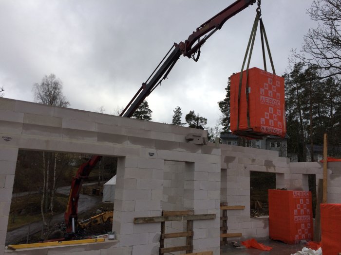 Kran lyfter en orange pall med Aeroc innerväggsblock över en vit muryta på ett bygge.