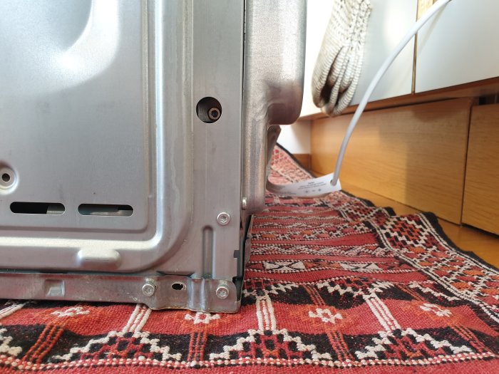 Bakre profil av en inbyggnadsugn med utstickande metallfästen, bredvid en vit elkabel på ett färgrikt tyg.