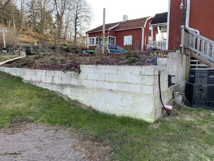 Betongmur i olika stadier av putsning med avloppsrör, intill röd stuga och trädgård.