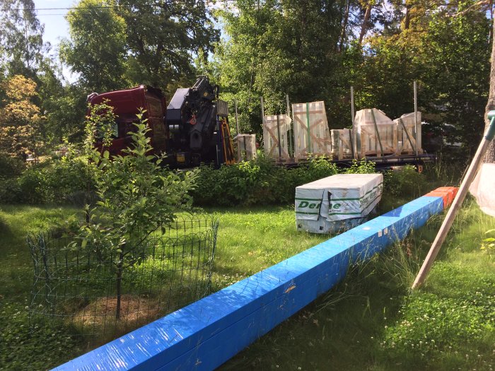 Leverans av byggmaterial som takstolar och fönster på en gräsmatta med skog i bakgrunden.