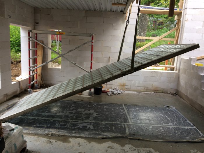 Stort härdad betongfönster som lyfts med rep inuti en byggnadsstomme, med en stege i bakgrunden.