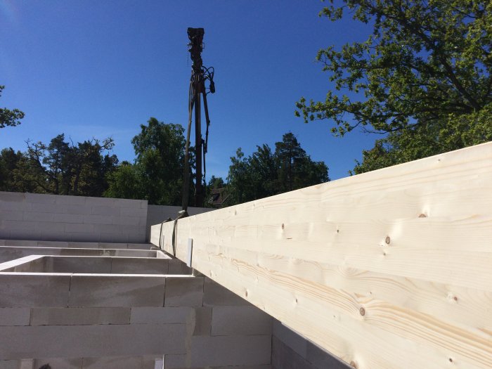 Kranarm ovanför nybyggd husstruktur med vita skumbetongblock och limträbalkar mot en klarblå himmel.