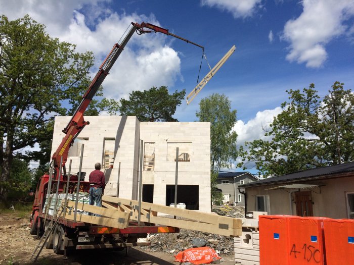 Byggarbetsplats med kranbil som lyfter en takstol mot en nybyggd husgrund med murrad balk och glasblocksfönster.