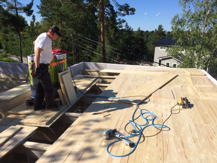 Person lägger råspont på tak med verktyg och snickarmaterial runtomkring under soligt väder.