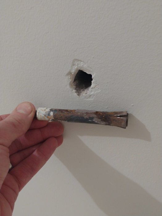 Hand som håller ett gammalt radiatorfäste framför ett hål i en vit lättbetongvägg där fästet lossnat.
