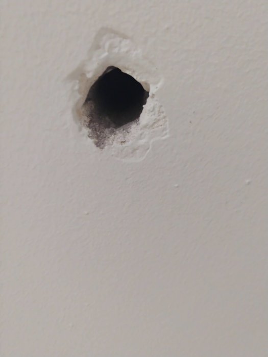 Vit vägg med ett ojämnt hål där ett radiatorfäste lossnat, synliga betongkanter och mörk bakgrund.