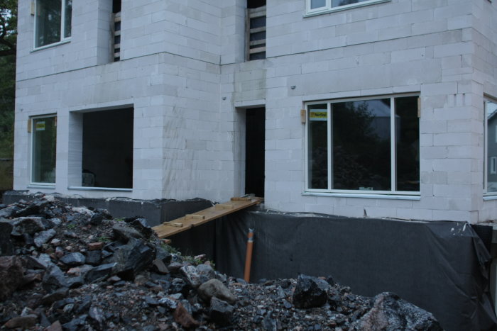 Nyinstallerade fönster i en vit murad byggnad med isolering synlig vid grunden, byggmaterial och grus i förgrunden.