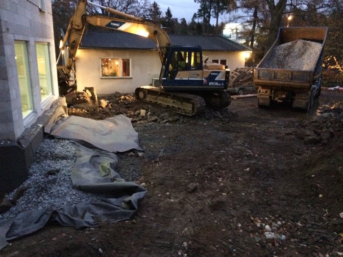 Återfyllning av grunden med grus och jordmassor vid ett hus, en grävmaskin och lastbil syns i arbete.