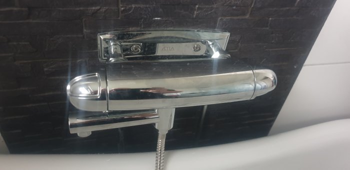 Kromad blandare monterad mot en mörk kakelvägg över ett badkar, med synliga vattenledningar anslutna undertill.