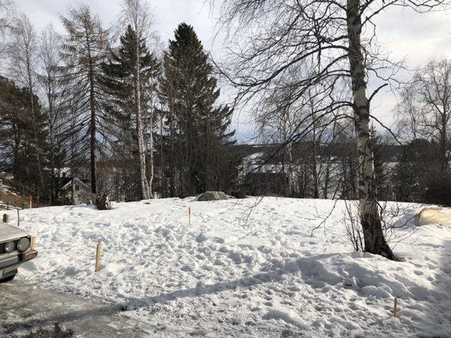 Tomt täckt med snö markerad för byggstart av garage, omgivet av träd och med en bil till vänster.
