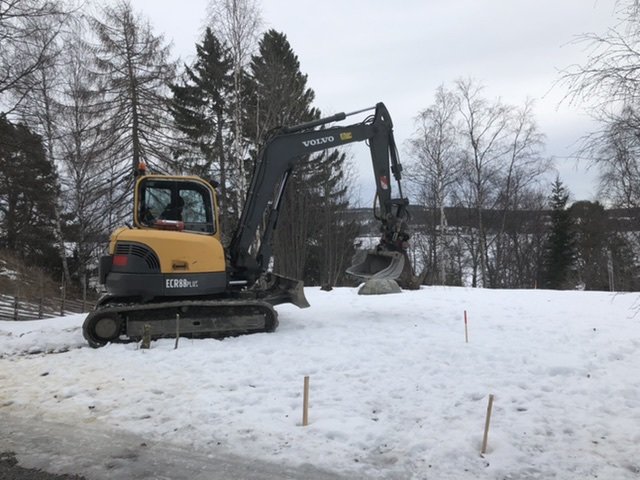 Grävmaskin på snöig tomt markerad med mätstickor inför garagebygge.
